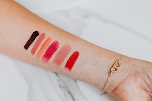 Close-up of various lipstick swatches on a woman's forearm with a heart bracelet.